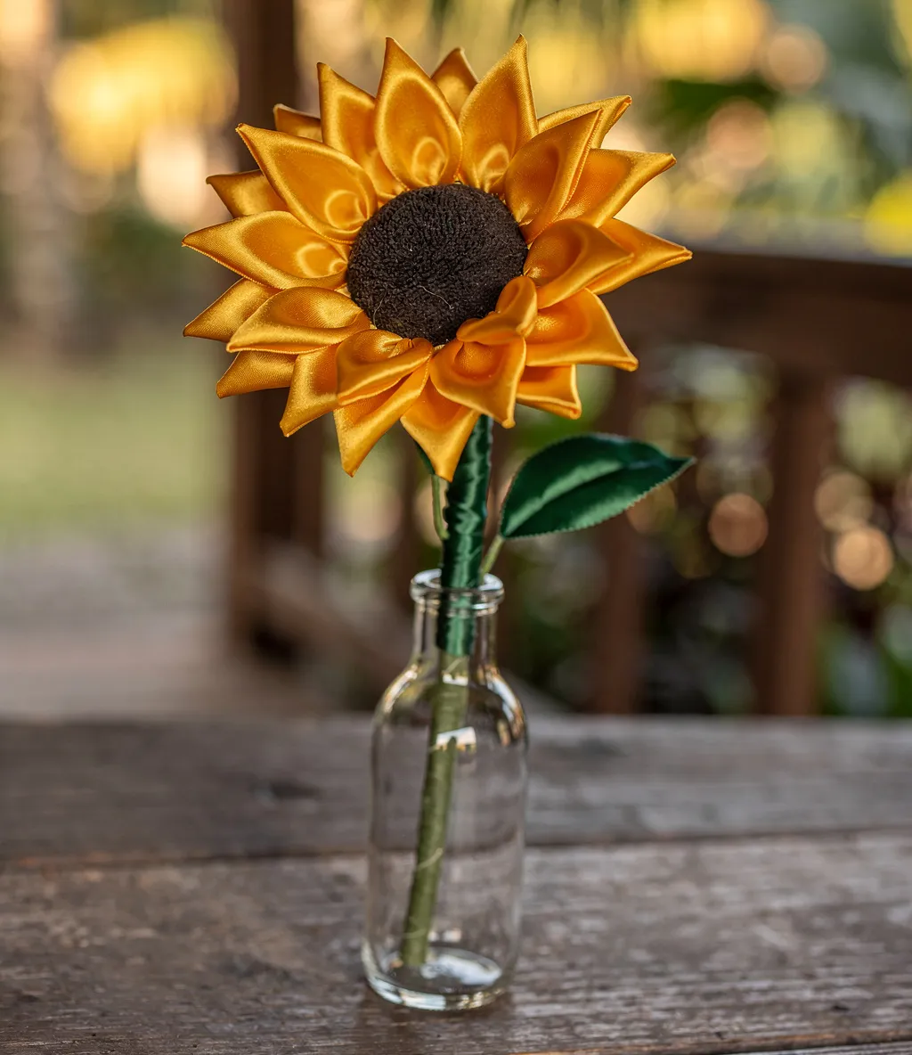 A single handmade satin sunflower with a brown center displayed in a small clear glass bottle.